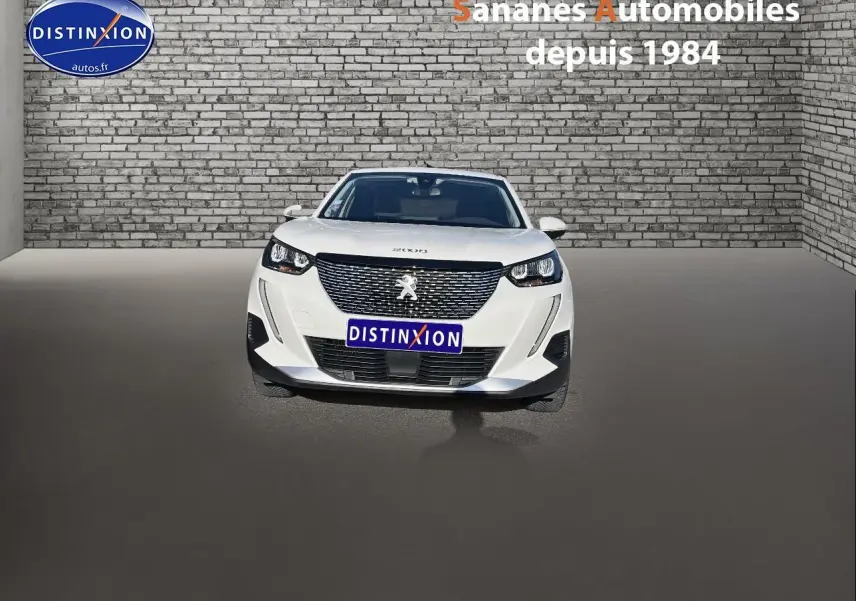 Vue frontale d'un Peugeot 2008 blanc 2020 avec calandre noire et phares LED, sur fond de mur en briques grises.