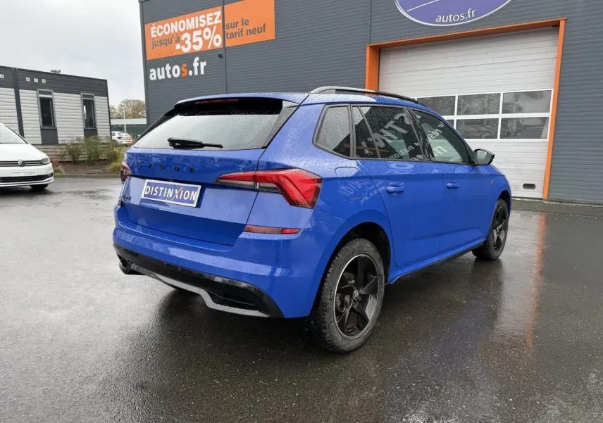 Vue 3/4 arrière droite d'un Skoda Kamiq bleu Energy avec jantes alliage noires et feux arrière LED sur parking humide