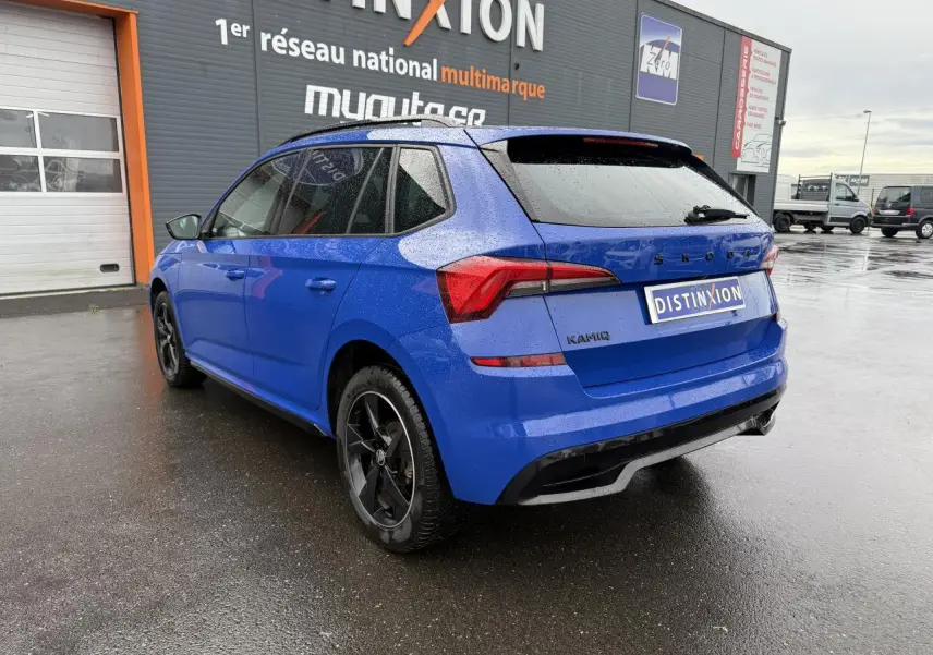 Vue 3/4 arrière gauche d'un Skoda Kamiq Bleu Energy avec jantes alliage noires et vitres surteintées sous la pluie.