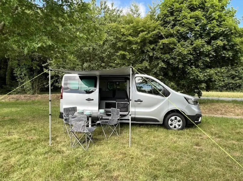 Renault Trafic Fourgon gris vu de profil droit avec auvent déployé et table de camping avec chaises sur herbe.