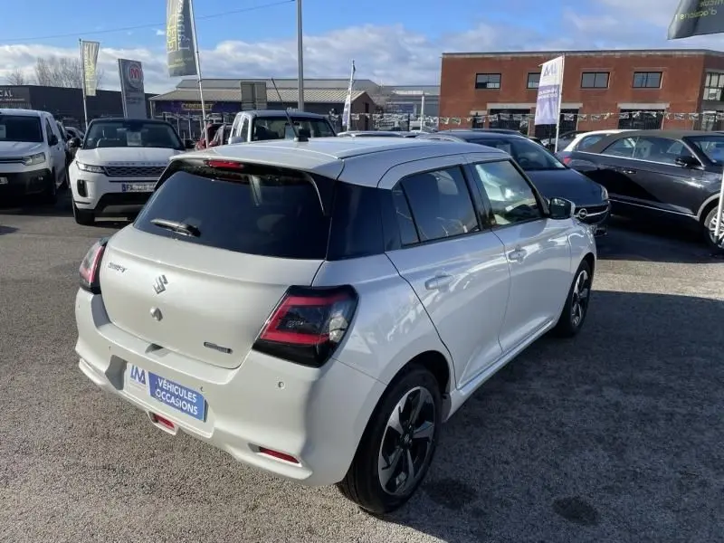 Vue 3/4 arrière droite d'une Suzuki Swift blanche 2025 hybride avec toit noir, garée sur un parking extérieur.