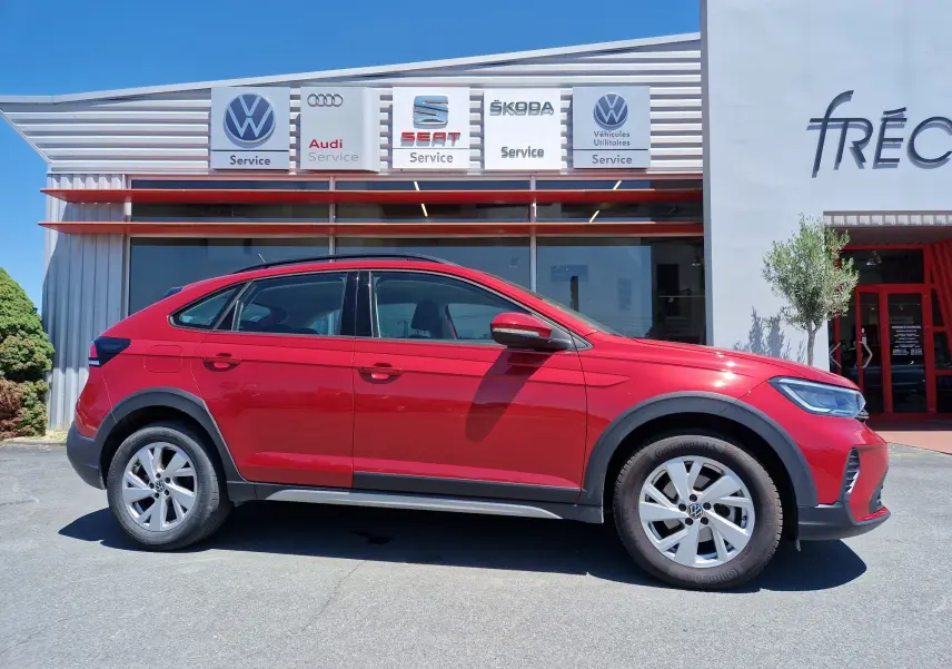 Vue de profil côté gauche d'un Volkswagen Taigo rouge roi garé devant un service auto multimarques.