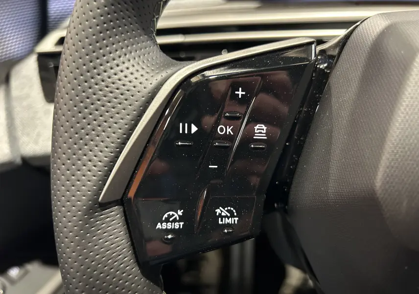 Gros plan sur les commandes gauche du volant en cuir perforé du Peugeot 5008 III Hybrid 145 GT, boutons multifonctions noirs.