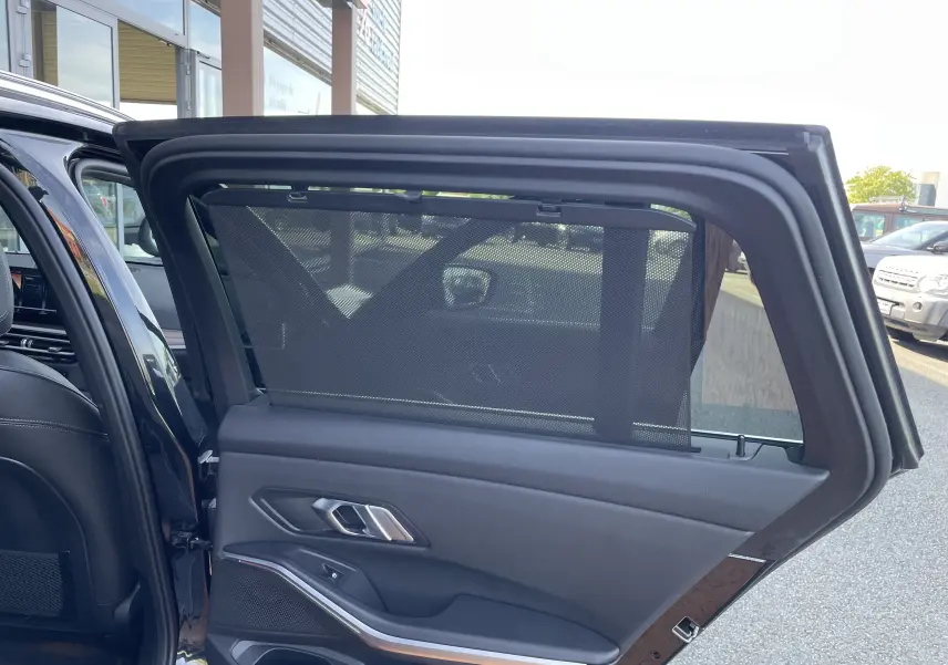Vue intérieure du côté gauche arrière de la BMW Série 3 Touring G21 noire, avec store pare-soleil déployé sur la vitre.
