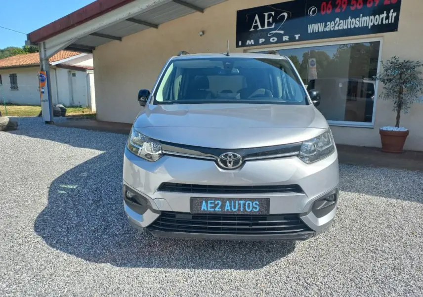 Vue frontale d'un Toyota Proace City Verso gris 2023, avec plaque AE2 AUTOS, stationné devant un bâtiment commercial.