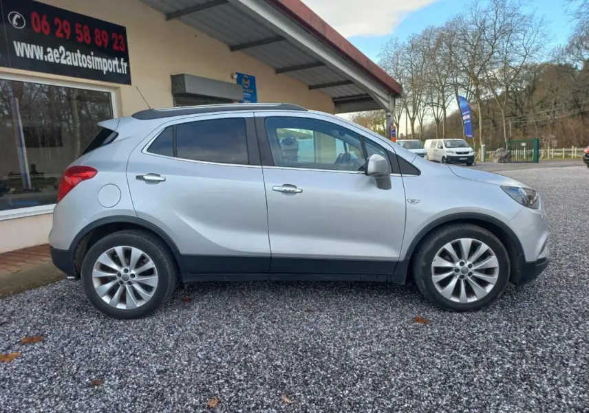 Opel Mokka X gris vue de profil côté gauche, avec jantes alliage et toit ouvrant, stationné devant un bâtiment.