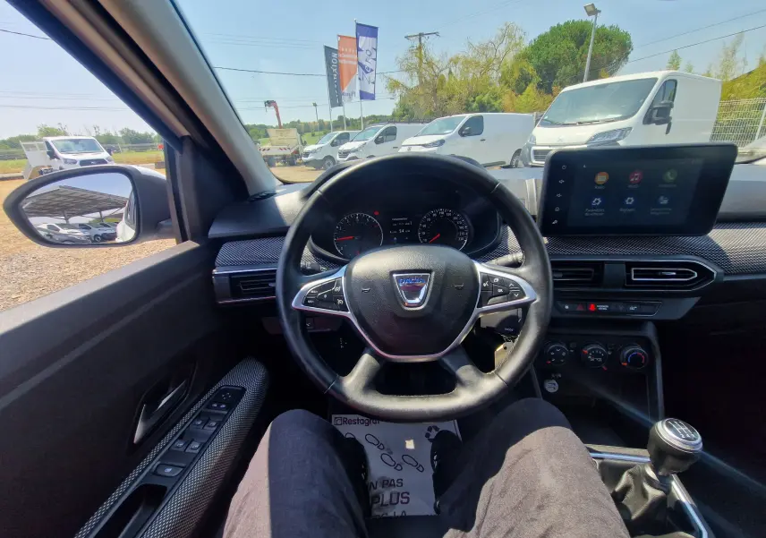 Vue intérieure centrée sur le volant Dacia Sandero blanc, tableau de bord et levier de vitesses manuelle visibles.