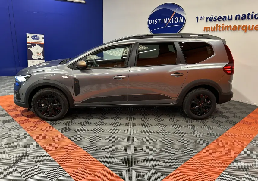 Vue de profil côté gauche d'un Dacia Jogger gris schiste métal avec jantes noires et barres de toit en intérieur showroom.