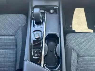 Vue plongeante sur la console centrale noire brillante du Volvo S60 2024 avec levier de vitesse et porte-gobelets entre sièges gris à motifs.
