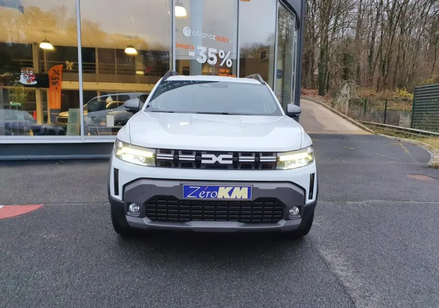 Vue frontale d’un Dacia Duster blanc glacier 2025 avec calandre noire et phares allumés devant un bâtiment vitré.