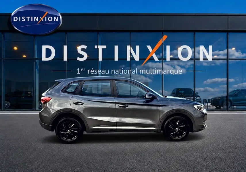 Profil droit du MG ZS Camden grey hybride 2026, avec jantes noires et lignes épurées devant un showroom Distinxion.
