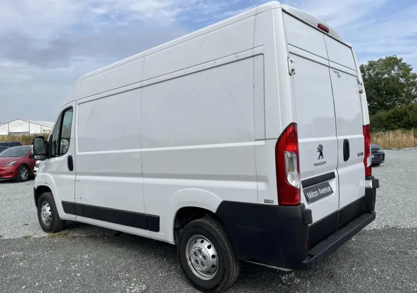 Vue 3/4 arrière droite d'un Peugeot Boxer fourgon blanc avec portes arrière battantes et protections noires sur un sol gravillonné.