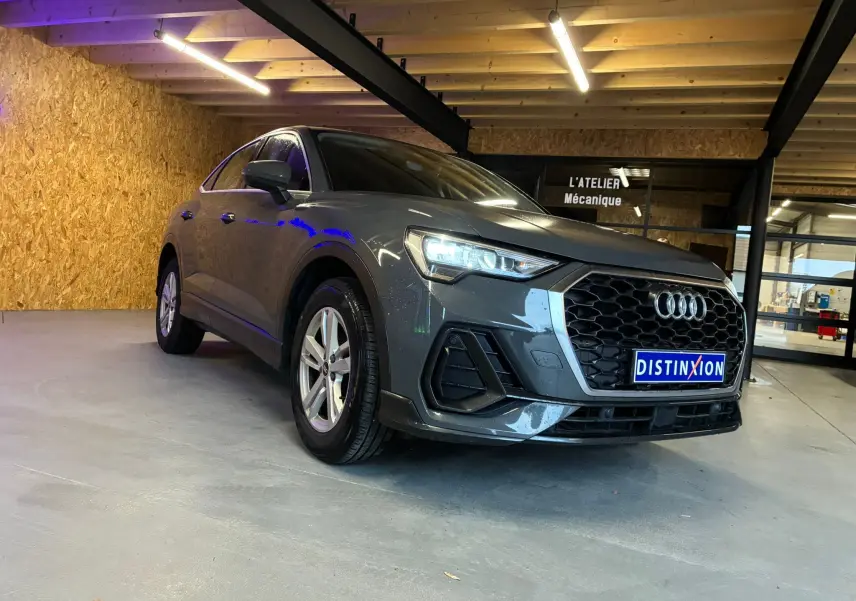 Audi Q3 Sportback gris en 3/4 avant droit, avec calandre noire hexagonale et jantes aluminium 17 pouces, dans un garage.