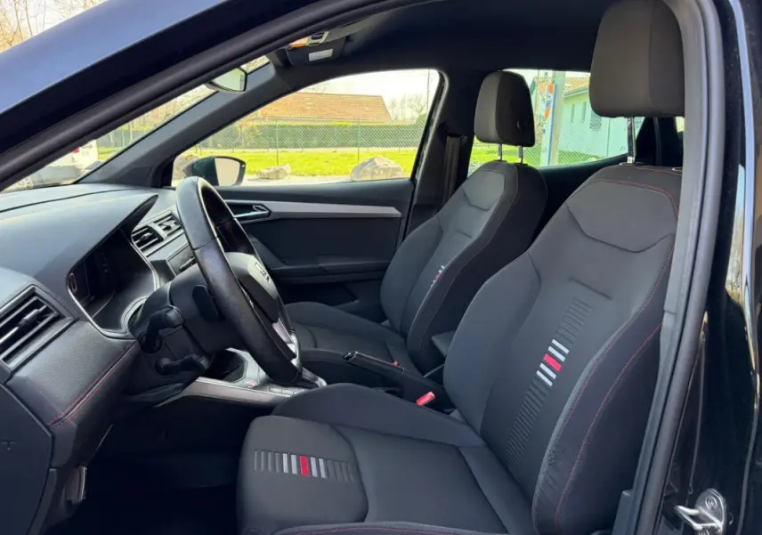 Vue intérieure côté conducteur du SEAT Arona noir 2020, mettant en valeur les sièges sport avec surpiqûres rouges et détails colorés.