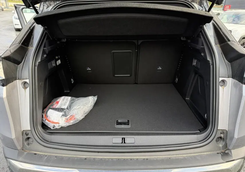 Coffre ouvert vu de l'arrière du Peugeot 3008 Hybrid gris, avec tapis noir et kit de dépannage visible à gauche.