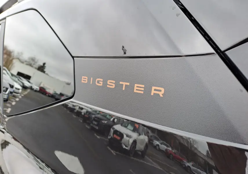 Gros plan sur l’inscription Bigster sur la carrosserie noir nacré d’un Dacia Bigster, reflet de voitures en arrière-plan.