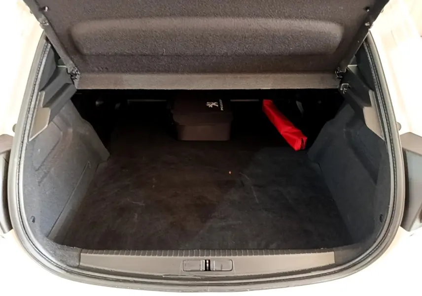 Vue intérieure du coffre ouvert d'une Peugeot 208 électrique blanche, avec tapis noir et accessoires rangés à l'arrière.