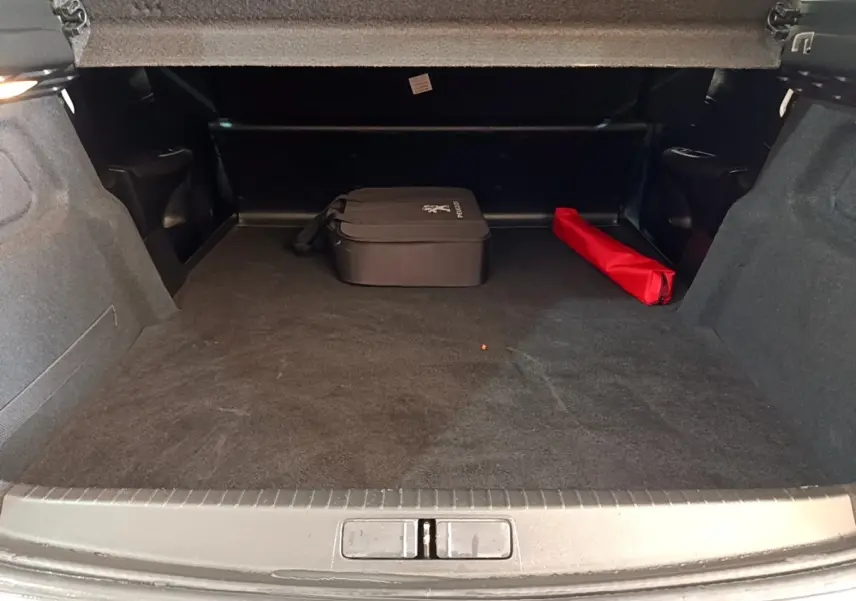 Coffre ouvert d’une Peugeot 208 électrique blanche, montrant un espace de chargement avec une trousse noire et un objet rouge.