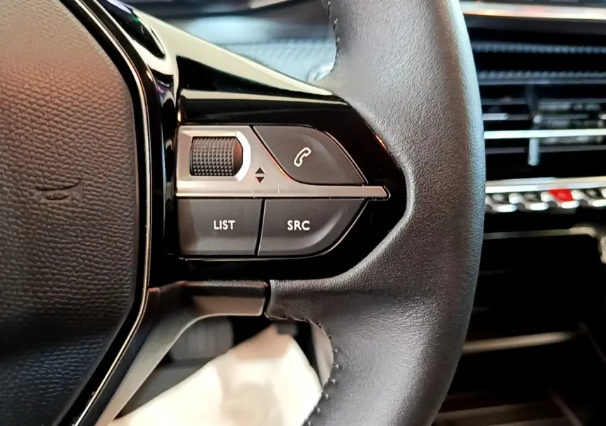 Gros plan sur les commandes gauche du volant cuir noir du Peugeot 208 électrique blanc, avec boutons de gestion audio et appel.