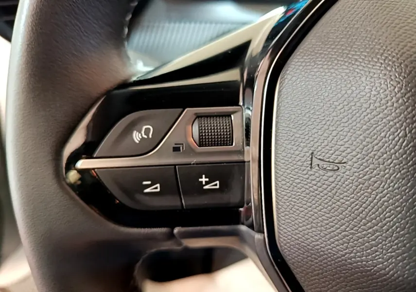 Gros plan sur les commandes gauche du volant cuir noir de la Peugeot 208 électrique blanche, avec boutons multifonctions.