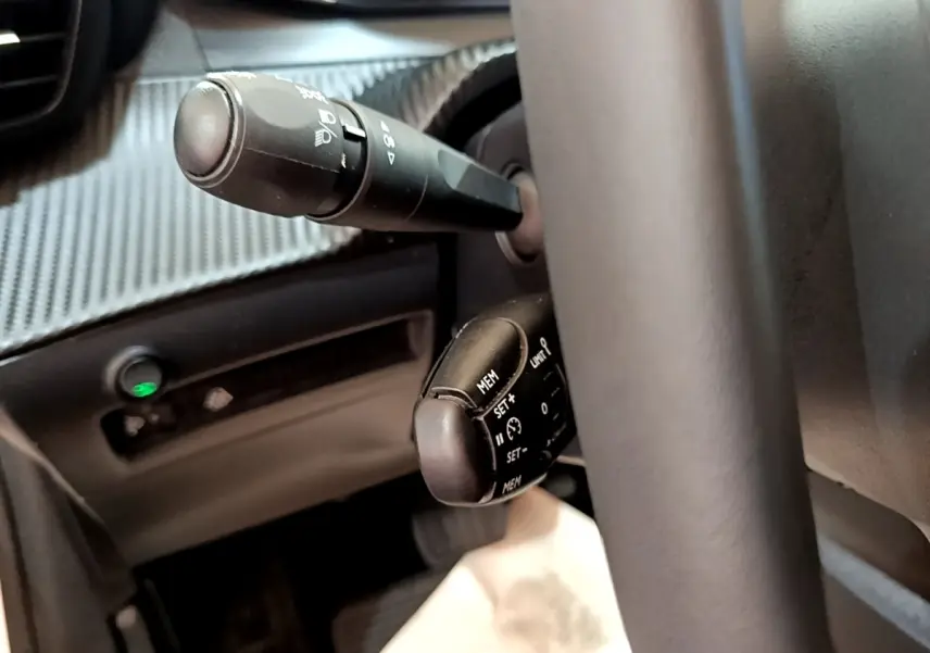 Gros plan sur les commandes au volant noir de la Peugeot 208 électrique blanche, avec levier de clignotants et régulateur de vitesse.