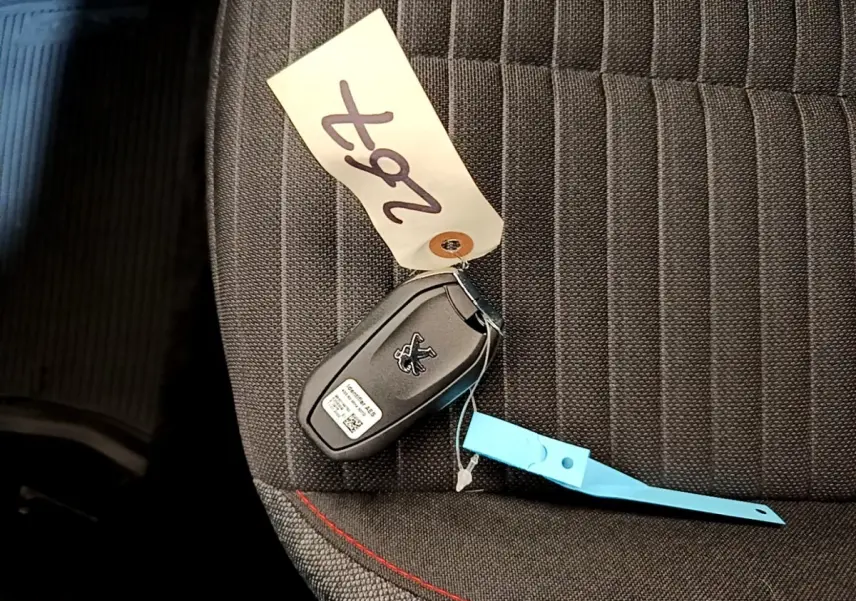 Clé de voiture Peugeot posée sur un siège tissu gris foncé avec surpiqûres rouges, intérieur visible partiellement.