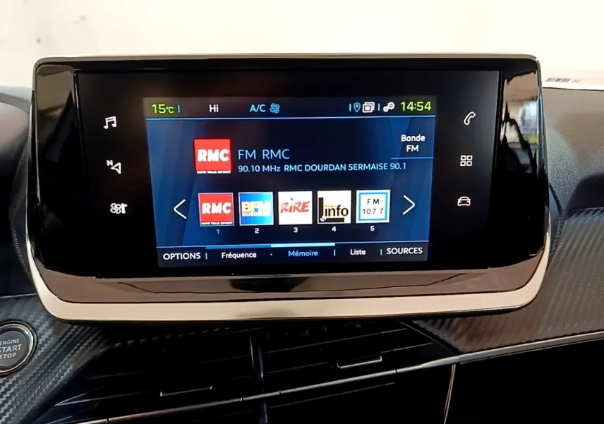 Écran tactile central affichant la radio FM dans l'habitacle d'une Peugeot 208 électrique blanche, vue de face.