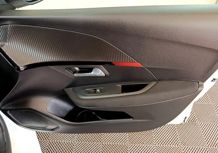Vue intérieure du panneau de porte côté conducteur de la Peugeot 208 électrique blanche, avec insert carbone et détail rouge.