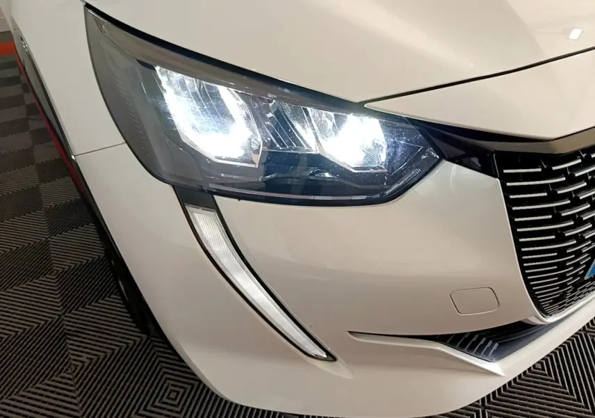Gros plan sur l'avant droit blanc d'une Peugeot 208 électrique 2022, mettant en valeur le phare LED et la calandre noire.