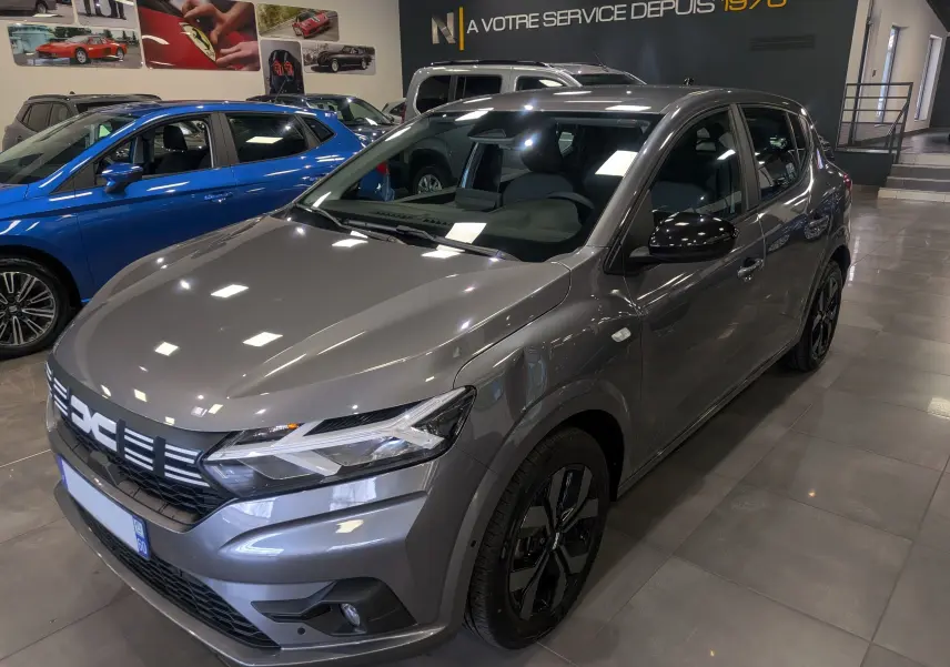 Vue 3/4 avant droite d'une Dacia Sandero TCe 90 gris schiste 2025 en showroom, avec jantes noires distinctives.