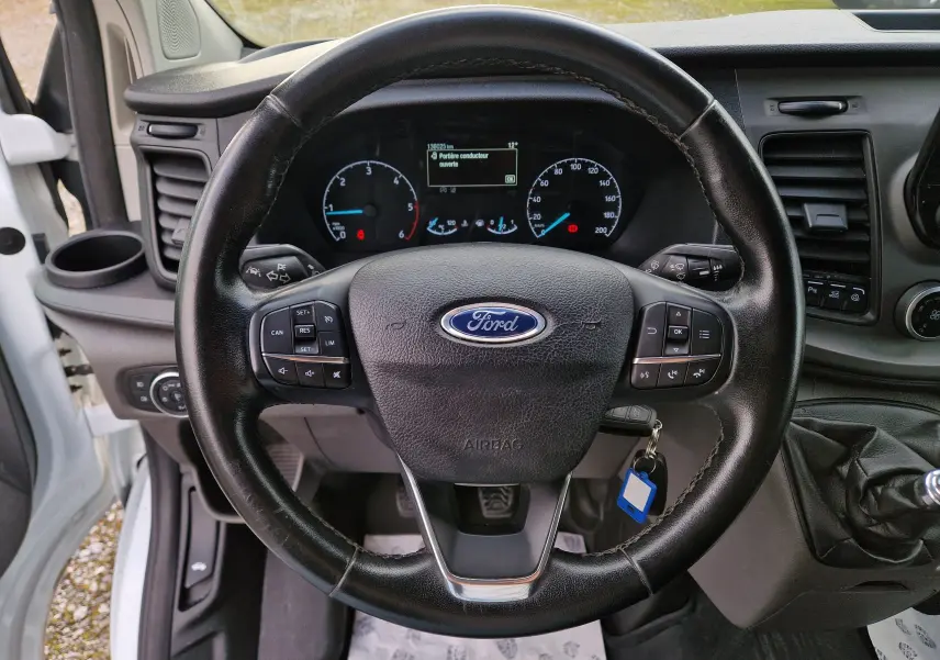 Vue intérieure centrée sur le volant noir du Ford Transit Custom Fourgon blanc, avec tableau de bord et levier de vitesses manuel visibles.