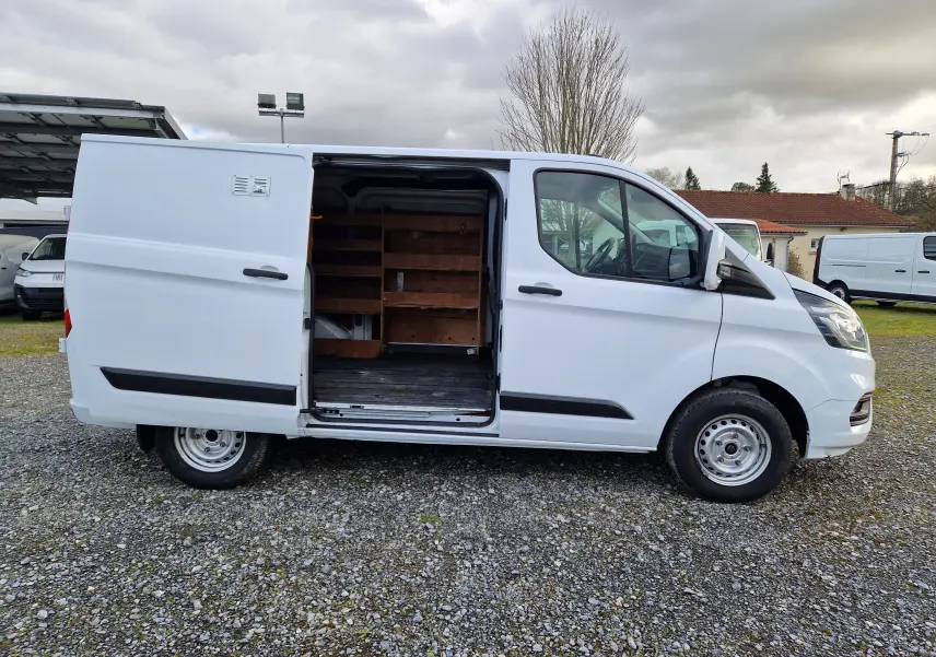 Ford Transit Custom Fourgon blanc, vue de profil côté gauche sur un sol gravillonné, sans fenêtres latérales arrière.