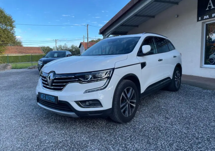 Renault Koleos blanc vu de 3/4 avant droit, avec calandre chromée et jantes alliage foncé, stationné devant un bâtiment.