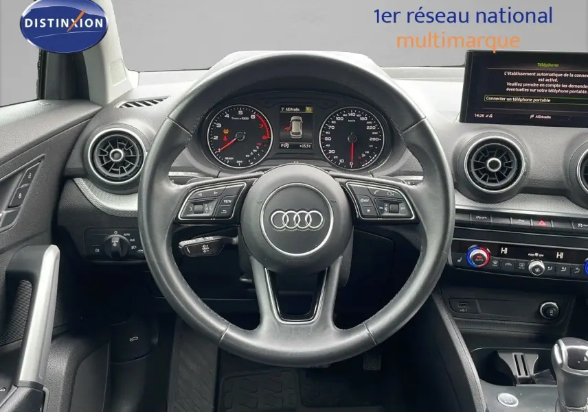 Vue intérieure centrée sur le volant Audi Q2 noir, tableau de bord avec compteurs et écran multimédia visible.