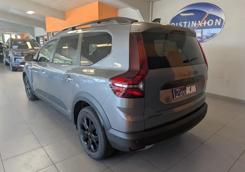 Vue 3/4 arrière droite du Dacia Jogger gris schiste métal avec jantes noires et toit équipé de barres de toit en showroom.