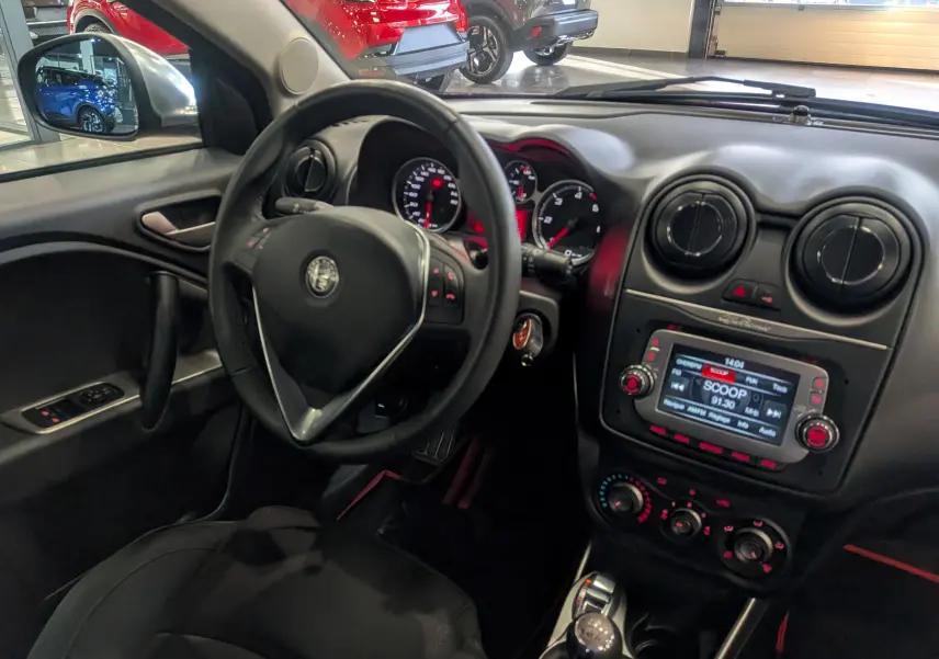 Vue intérieure côté conducteur de l'Alfa Romeo MiTo rouge, montrant le volant, tableau de bord et console centrale avec écran radio.
