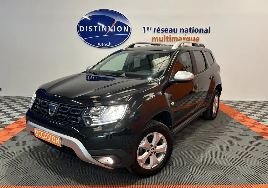 Dacia Duster noir en 3/4 avant droit, avec jantes alliage, barres de toit argentées et antibrouillards allumés.