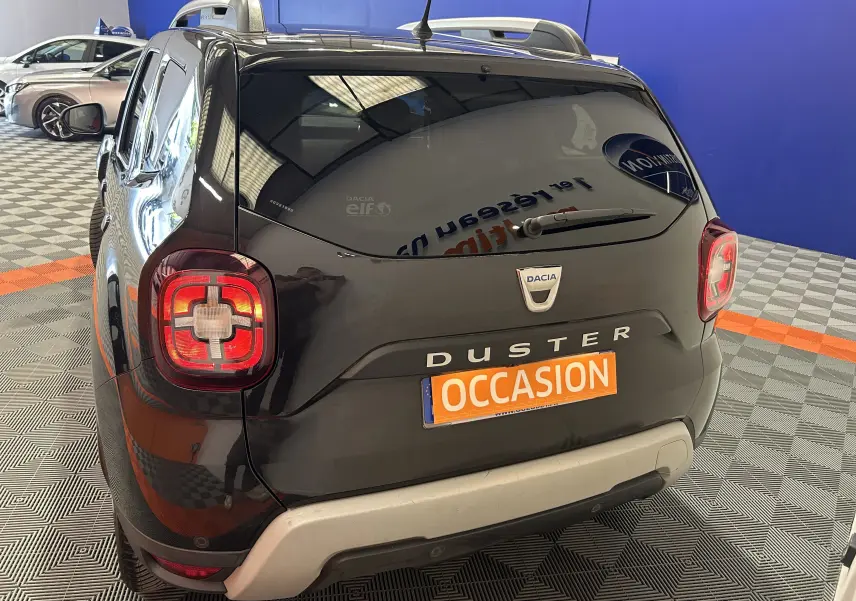 Vue arrière droite d'un Dacia Duster noir avec feux arrière allumés et plaque "OCCASION" en intérieur showroom.
