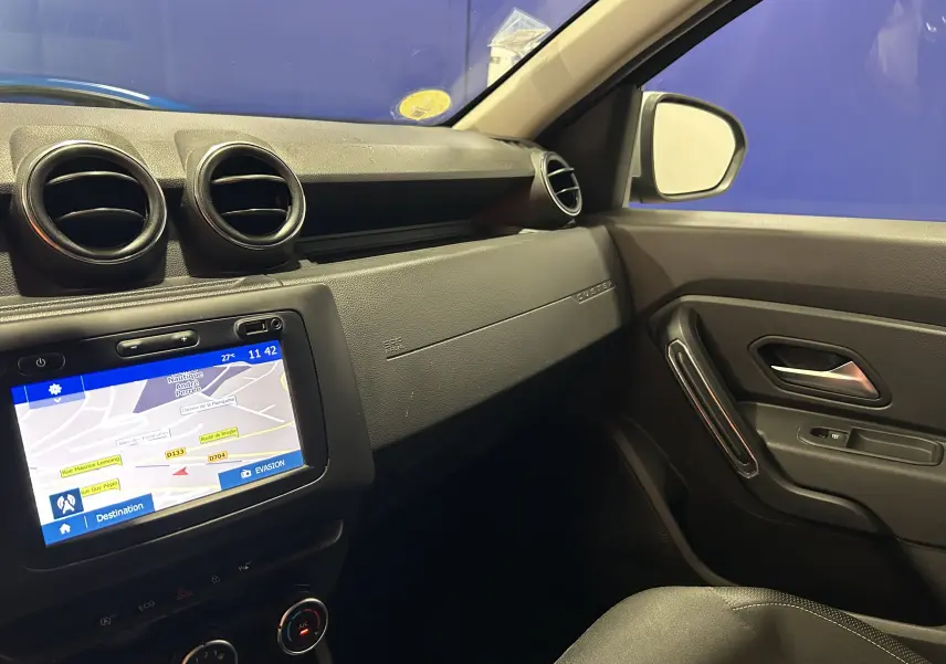 Vue intérieure côté passager du tableau de bord noir du Dacia Duster 2018 avec écran GPS allumé et aérateurs ronds.
