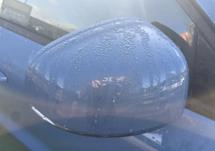 Rétroviseur côté gauche bleu Fjord Blue métallisé du Volvo XC40 2024 avec gouttes d'eau visibles.