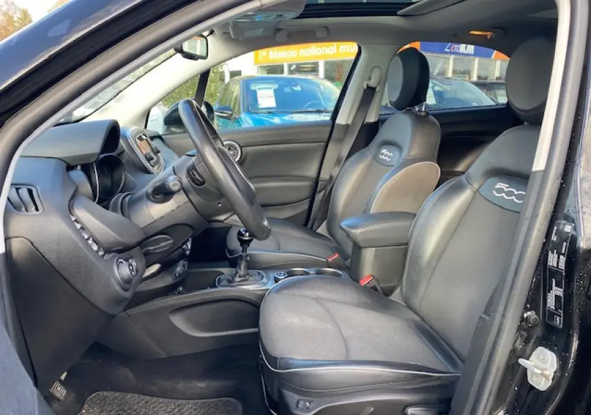 Intérieur noir vu côté conducteur d'une Fiat 500X 2016, sièges cuir avec logo 500 et toit panoramique ouvert.