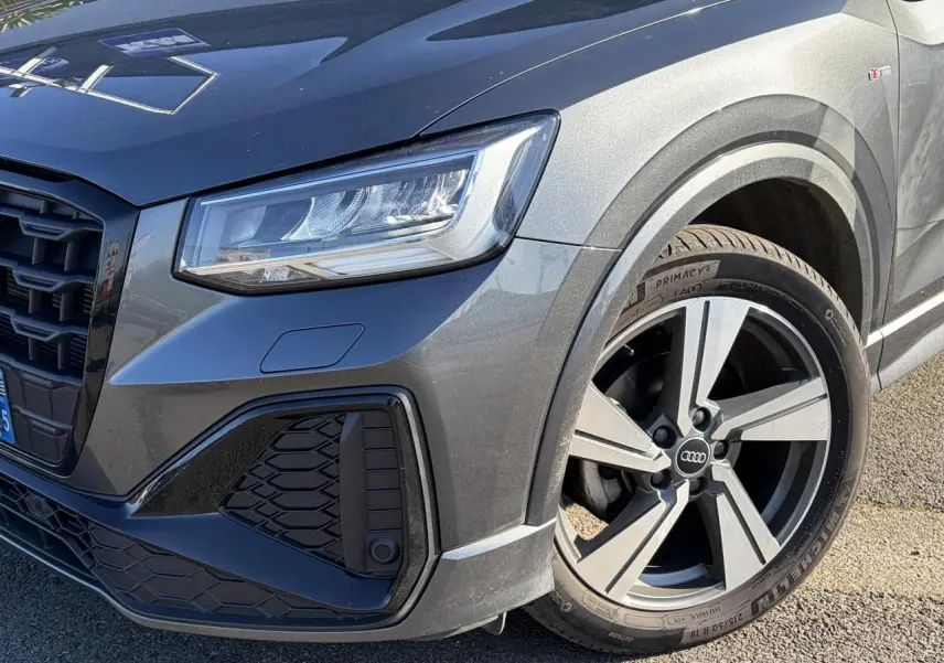 Gros plan sur l’avant droit de l’Audi Q2 gris Daytona 2024, mettant en valeur le phare LED et la jante alliage 18 pouces S line.