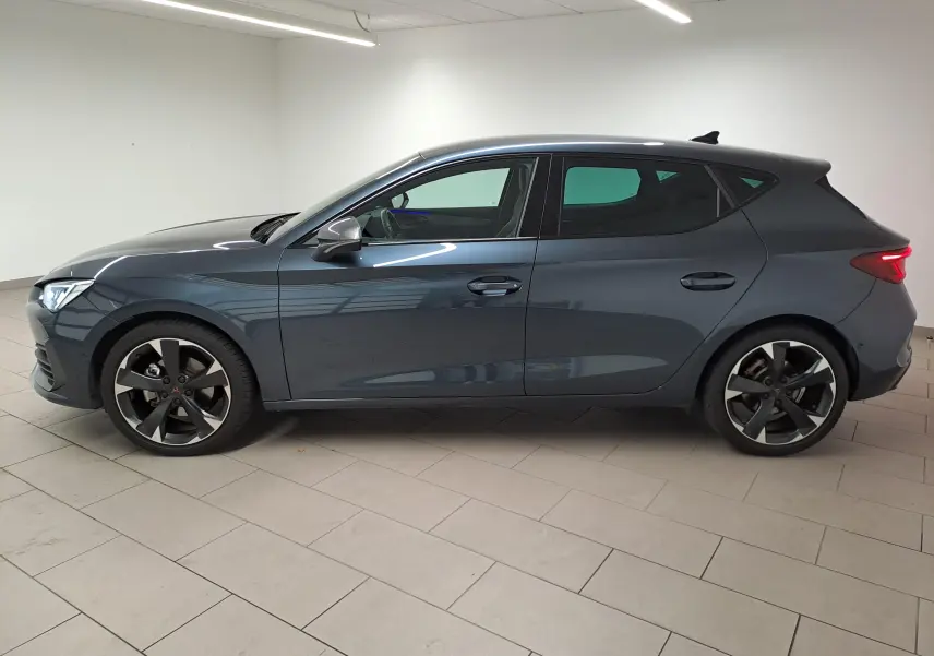 Profil côté gauche d'une CUPRA Leon 1.5 eTSI 150 ch gris magnétique 2023 avec jantes noires distinctives.