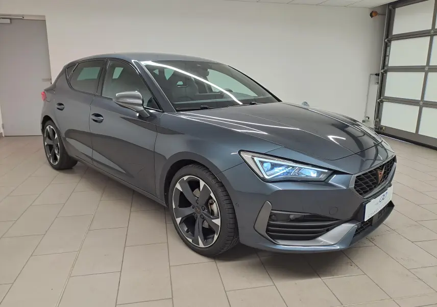 Vue 3/4 avant droit d'une CUPRA Leon 1.5 eTSI 150 ch gris magnétique avec jantes noires et phares LED allumés.