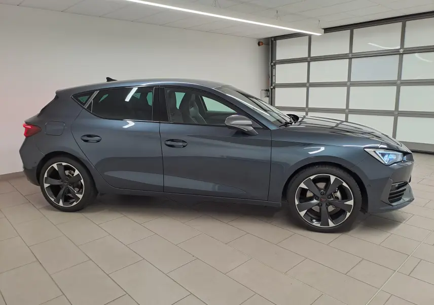 Vue de profil côté gauche d'une CUPRA Leon 1.5 eTSI 150 ch gris magnétique avec jantes noires et grises dans un garage.