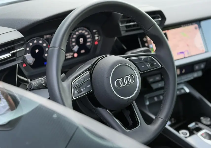 Vue rapprochée du volant cuir noir multifonctions d'une Audi A3 Sportback 2024 avec tableau de bord numérique et écran tactile.