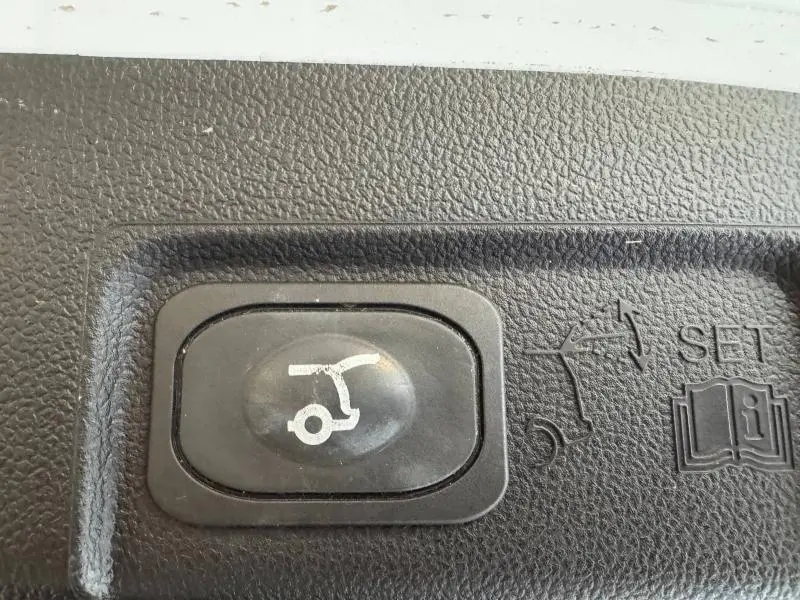 Bouton noir d'ouverture électrique du hayon sur une Ford Puma blanc glacier, avec pictogrammes en relief sur fond texturé.