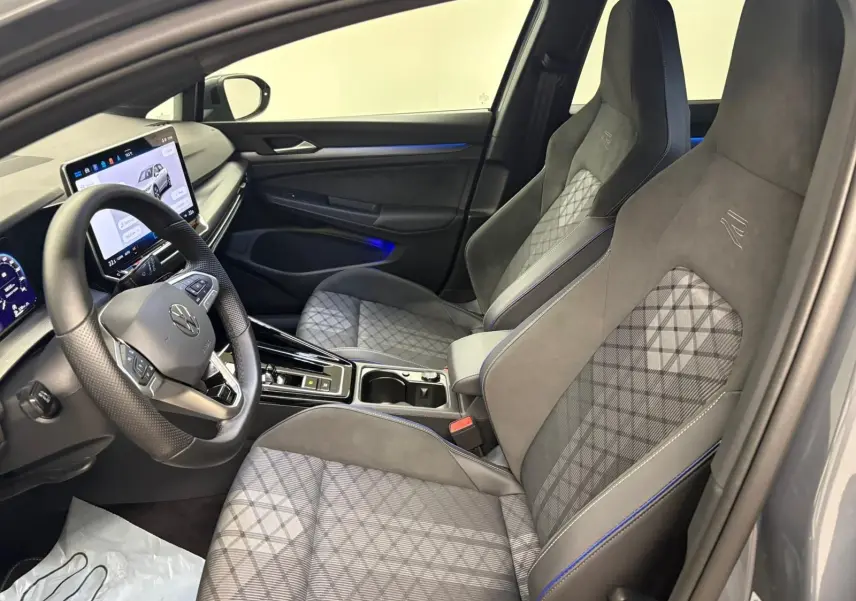 Vue intérieure côté conducteur de la Volkswagen Golf 1.5 eTSI R-Line 2025 avec sièges gris à surpiqûres bleues et écran tactile.