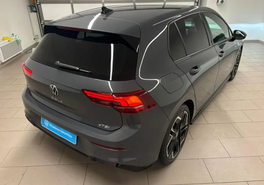 Vue 3/4 arrière droite d'une Volkswagen Golf 1.5 eTSI gris dauphin avec feux arrière allumés et jantes noires R-Line.