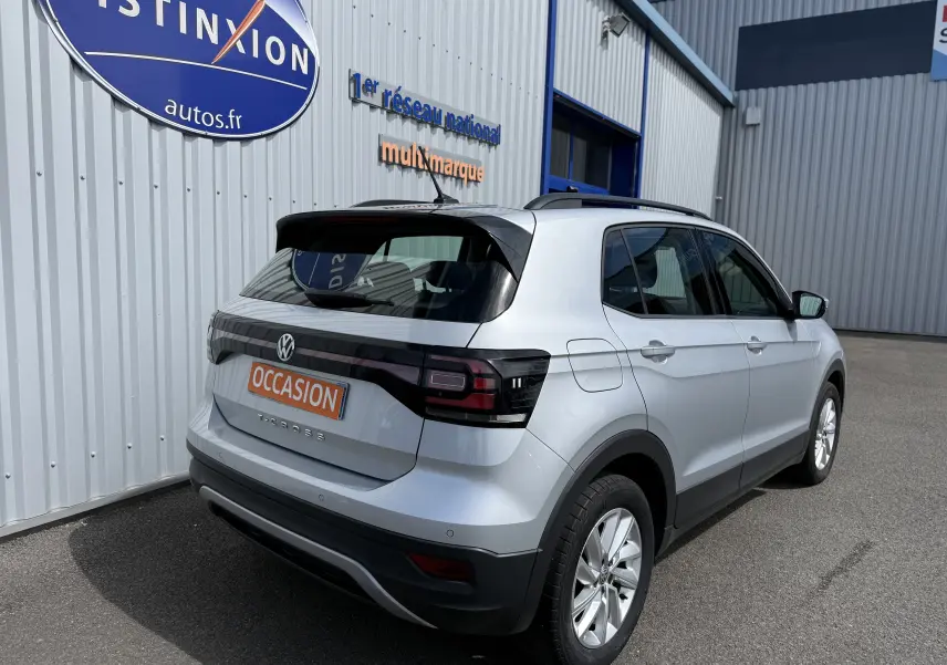 Vue 3/4 arrière droite d'un Volkswagen T-Cross gris clair avec feux arrière noirs et toit noir, stationné devant un bâtiment.